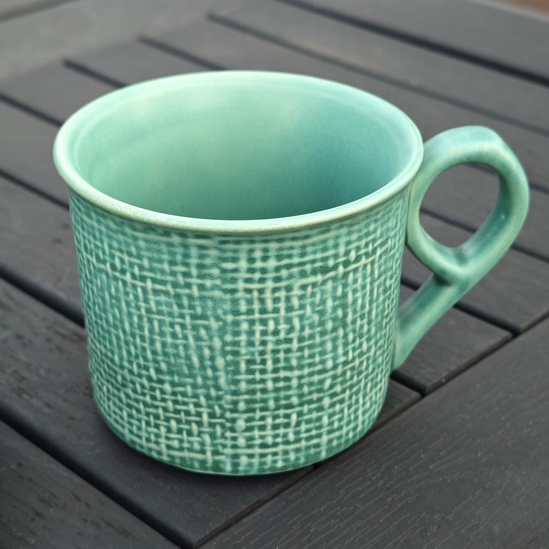 May include: A teal ceramic mug with a textured woven pattern. The mug has a rounded handle and a smooth rim. The mug is sitting on a dark wooden surface.
