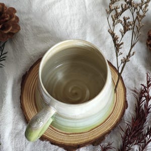 May include: A handmade ceramic mug with a wide, round shape and a single handle. The mug features a gradient of colors, transitioning from white at the top to light green at the base. It sits on a round wooden coaster, with a neutral background.