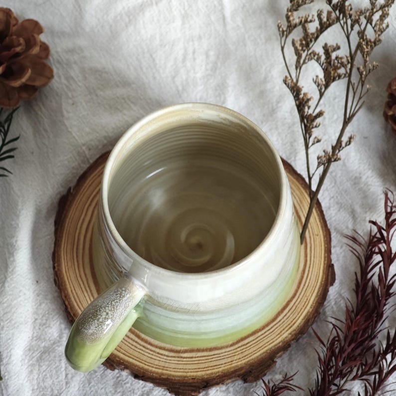 May include: A handmade ceramic mug with a wide, round shape and a single handle. The mug features a gradient of colors, transitioning from white at the top to light green at the base. It sits on a round wooden coaster, with a neutral background.