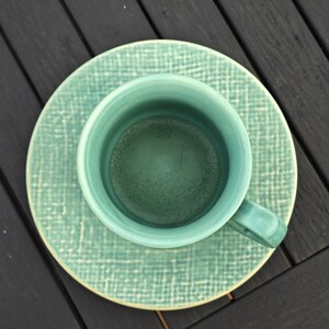 May include: An overhead shot of a teal ceramic teacup and saucer set. The cup has a handle and sits on a matching saucer. Both pieces have a textured, crosshatch pattern. The set is a solid teal color.