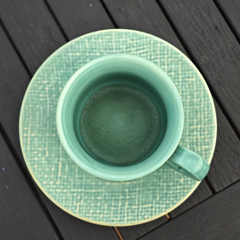 May include: An overhead shot of a teal ceramic teacup and saucer set. The cup has a handle and sits on a matching saucer. Both pieces have a textured, crosshatch pattern. The set is a solid teal color.