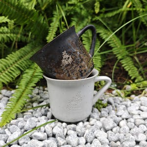 May include: Two ceramic mugs, one stacked on the other. The top mug is dark brown with a textured finish and a white graphic. The bottom mug is light grey with a white graphic and the text "Yu Garden Studio".