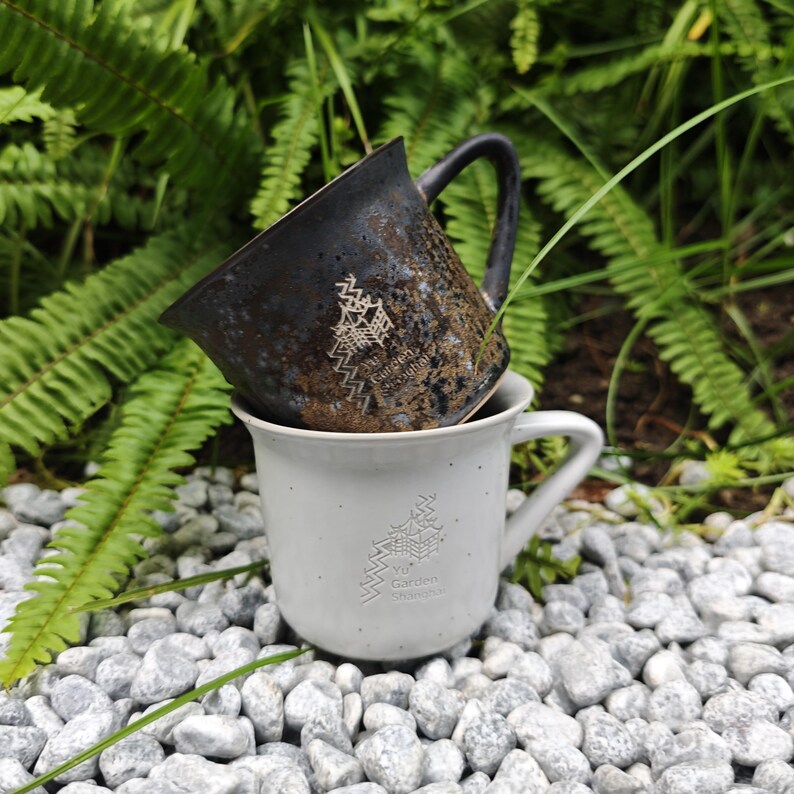 May include: Two ceramic mugs, one stacked on the other. The top mug is dark brown with a textured finish and a white graphic. The bottom mug is light grey with a white graphic and the text "Yu Garden Studio".