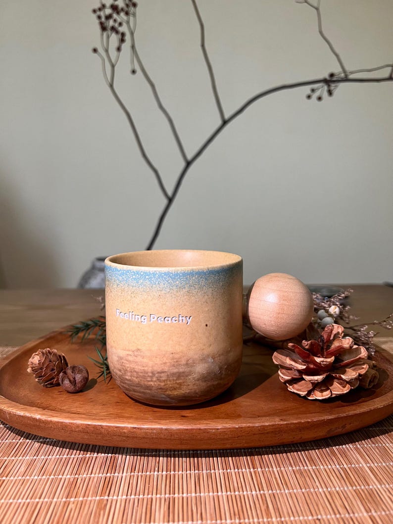 May include: A ceramic mug with a gradient design, transitioning from a light blue rim to a tan and brown base. The mug is on a wooden tray with a wooden ball, pinecone, and sprigs. The text "Feeling Peachy" is on the mug.