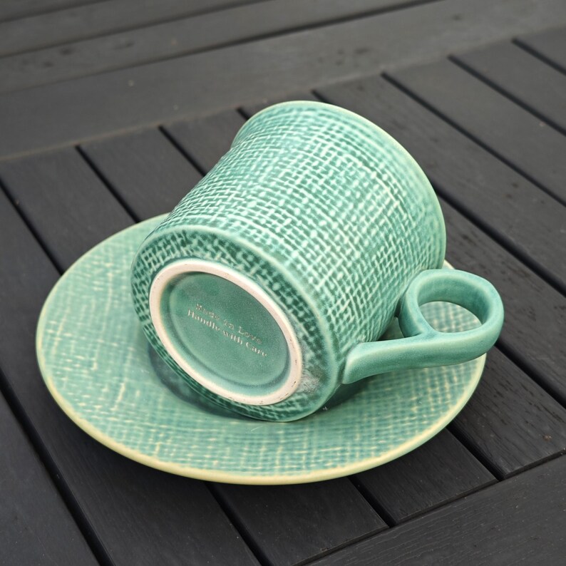 May include: A teal ceramic teacup and saucer set. The cup has a rounded handle and a textured, woven pattern. The saucer is round with a matching texture. The bottom of the cup reads "Made with Love, Handle with Care."