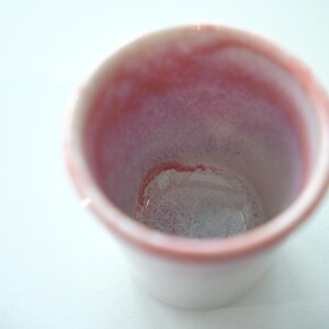 May include: A small, white ceramic cup with a pink interior and rim. The inside of the cup has a gradient of pink and purple hues. The cup is empty and sits on a white surface.
