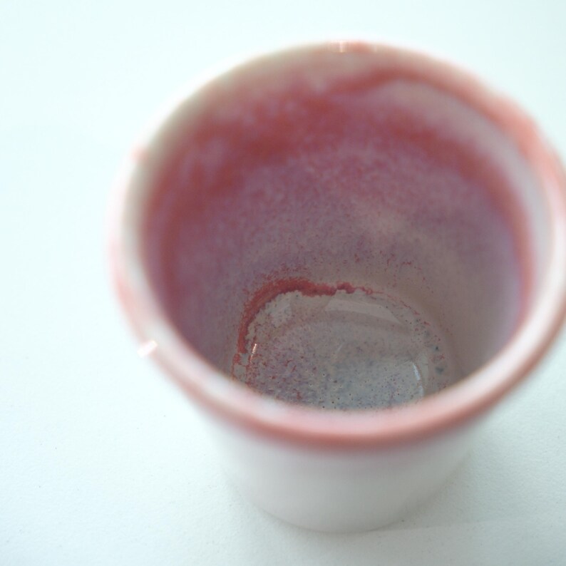 May include: A small, white ceramic cup with a pink interior and rim. The inside of the cup has a gradient of pink and purple hues. The cup is empty and sits on a white surface.