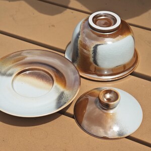 May include: A ceramic tea set consisting of an inverted bowl, a matching saucer, and a lid. The pieces feature a glazed finish with blended gradients of white, brown, and charcoal grey. The bowl is positioned upside down, revealing its base.