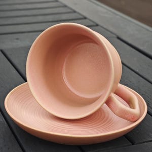 May include: A pink ceramic teacup and saucer set. The cup has a curved handle and sits atop a matching saucer. The saucer features concentric circle detailing. The set is a soft, muted pink color.