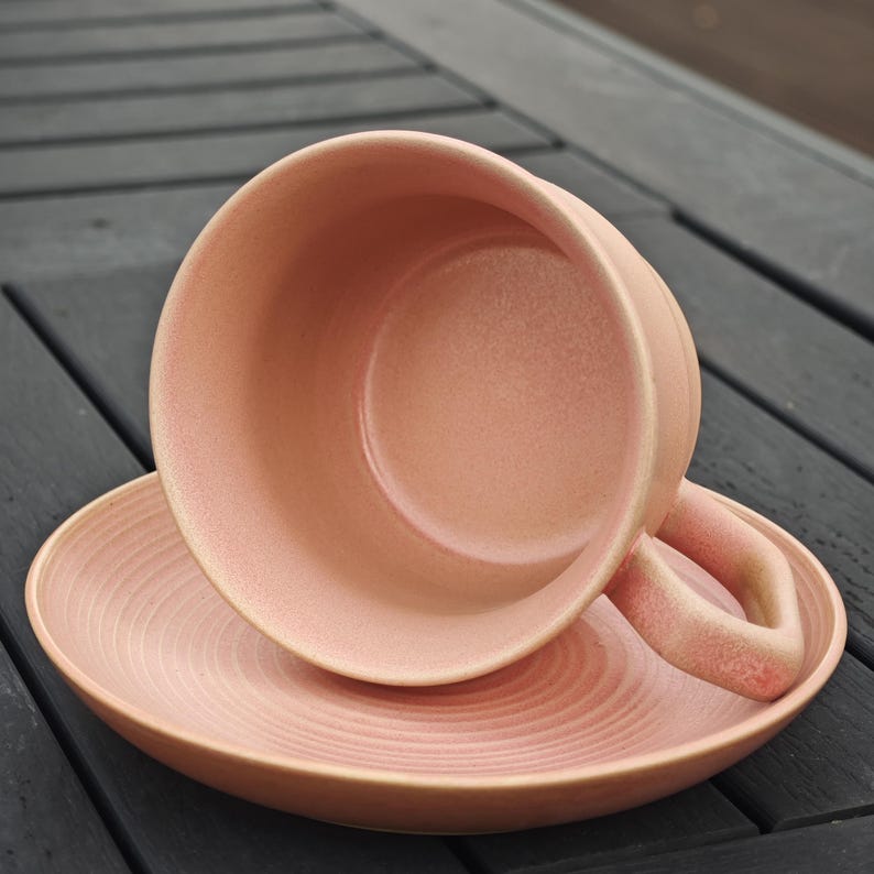 May include: A pink ceramic teacup and saucer set. The cup has a curved handle and sits atop a matching saucer. The saucer features concentric circle detailing. The set is a soft, muted pink color.
