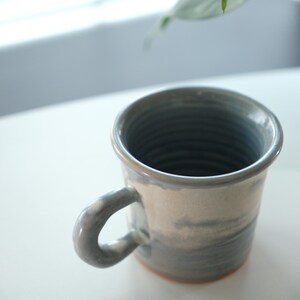 May include: A ceramic mug with a grey and white marbled pattern. The mug features a curved handle and a slightly flared rim. The interior is a darker grey, and the base has a reddish-brown colour. Suitable for hot or cold drinks.