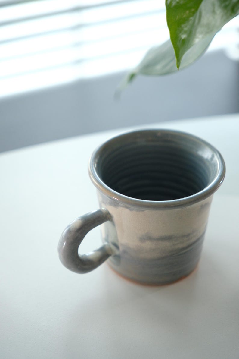 May include: A ceramic mug with a grey and white marbled pattern. The mug features a curved handle and a slightly flared rim. The interior is a darker grey, and the base has a reddish-brown colour. Suitable for hot or cold drinks.
