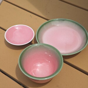 Puede incluir: Un juego de tres piezas de cer&aacute;mica que incluye un platillo rosa peque&ntilde;o, un plato rosa m&aacute;s grande con borde verde y un cuenco rosa profundo con borde verde. Cada pieza presenta un acabado liso y brillante con un degradado de rosa a verde.