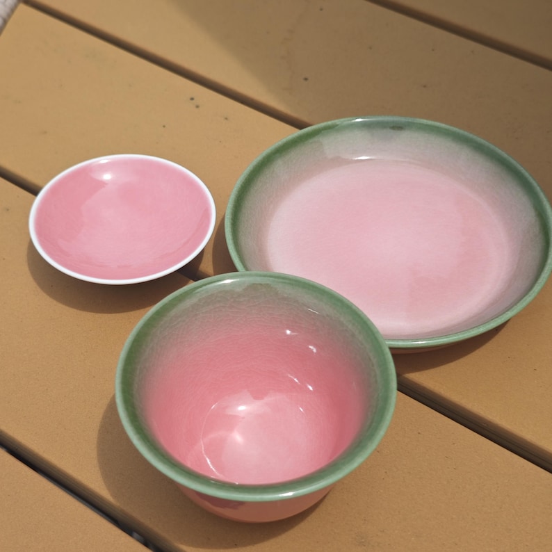 Puede incluir: Un juego de tres piezas de cer&aacute;mica que incluye un platillo rosa peque&ntilde;o, un plato rosa m&aacute;s grande con borde verde y un cuenco rosa profundo con borde verde. Cada pieza presenta un acabado liso y brillante con un degradado de rosa a verde.