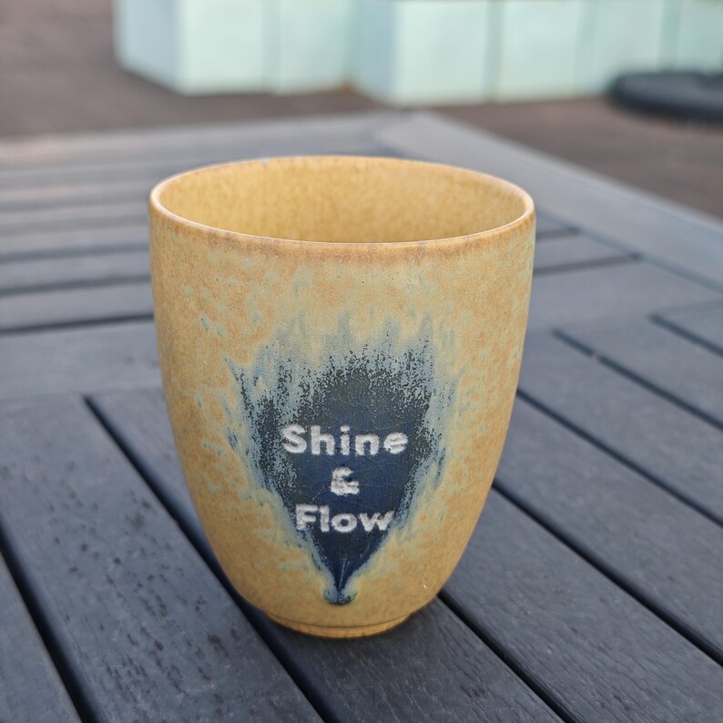Puede incluir: Un vaso de cer&aacute;mica con exterior beige e interior azul oscuro. El vaso presenta las palabras "Shine & Flow" en letras blancas. La taza mide aproximadamente 10 cm de alto y tiene una forma suave y redondeada.
