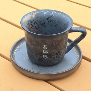 May include: A speckled, dark gray ceramic teacup and saucer set. The cup has a handle and the saucer is round. The cup and saucer have a speckled pattern with gold accents. The cup has the text "萬瑞緣" in white.