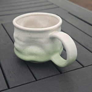 Op de afbeelding: Een handgemaakte keramische mok met een wit-groene ombre glazuur. De mok heeft een afgeronde body, een grote handgreep en een gespikkelde textuur. De mok is geschikt voor koffie, thee of andere dranken.