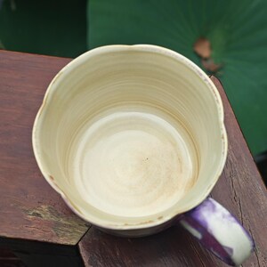 May include: A handmade ceramic mug with a unique, wavy rim. The interior is a creamy beige, while the exterior has a light beige hue with a purple handle. The mug is set on a wooden surface, suggesting a rustic aesthetic.