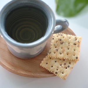 May include: A pale blue ceramic mug with a handle rests on a small, round wooden tray. Two square crackers with dark specks are beside the mug. The mug has a textured interior and a light blue exterior.