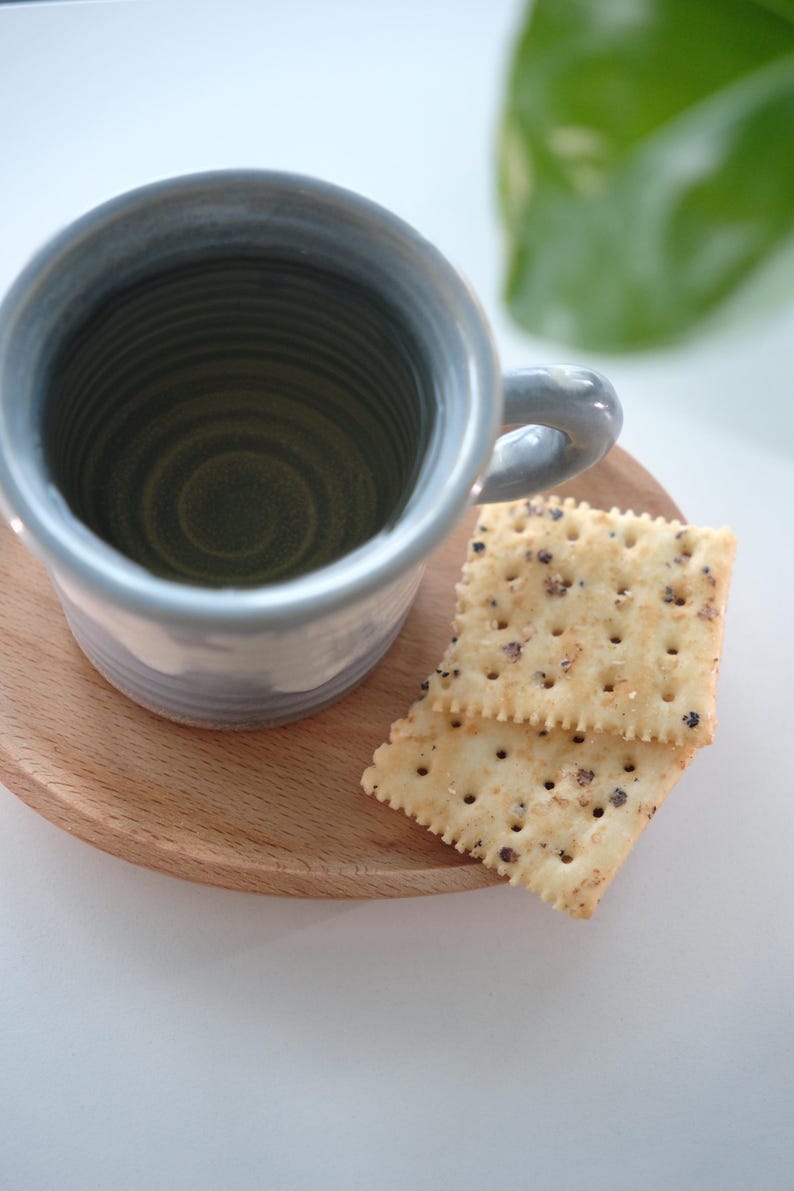 May include: A pale blue ceramic mug with a handle rests on a small, round wooden tray. Two square crackers with dark specks are beside the mug. The mug has a textured interior and a light blue exterior.