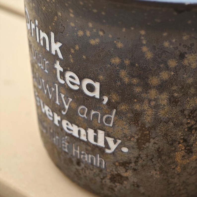 May include: Close-up of a dark brown ceramic mug with white text that reads "Drink your tea, slowly and reverently." The mug has a textured surface with small, star-like gold accents. The bottom of the mug has the name "Hanh" in white text.