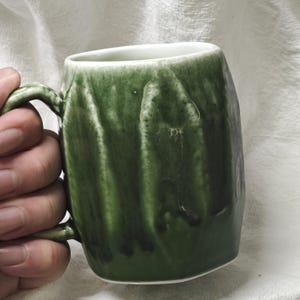 May include: A handmade ceramic mug with a dark green glaze. The mug has a unique, textured surface and a curved handle. The interior is white. The mug is held in a person's hand.