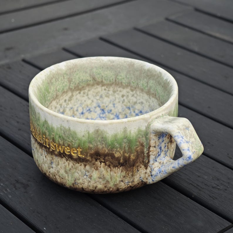 May include: A ceramic mug with a unique, textured design. The mug features a color palette of green, brown, and blue, with the words "make it sweet" in gold lettering. The handle has a distinctive angular shape.