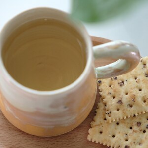 May include: A handmade ceramic mug with a light yellow and peach gradient, filled with a light-colored liquid. The mug has a rounded handle and sits on a wooden surface next to two crackers.