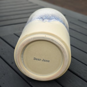 May include: A ceramic tumbler with a cream-colored base and a blue and white speckled glaze at the top. The bottom of the tumbler features the text Dear Jane stamped in dark lettering on the unglazed ceramic surface of the base.