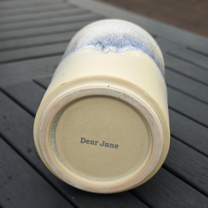 May include: A ceramic tumbler with a cream-colored base and a blue and white speckled glaze at the top. The bottom of the tumbler features the text Dear Jane stamped in dark lettering on the unglazed ceramic surface of the base.