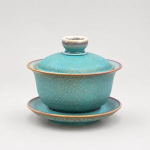 K&ouml;nnte beinhalten: Ein t&uuml;rkisfarbenes Gaiwan-Teeservice aus Keramik, bestehend aus einer Schale, einem Deckel mit Knaufgriff und einer passenden Untertasse. Die Glasur hat ein gesprenkeltes, strukturiertes Aussehen mit dezenten braunen R&auml;ndern.