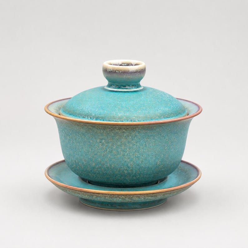 K&ouml;nnte beinhalten: Ein t&uuml;rkisfarbenes Gaiwan-Teeservice aus Keramik, bestehend aus einer Schale, einem Deckel mit Knaufgriff und einer passenden Untertasse. Die Glasur hat ein gesprenkeltes, strukturiertes Aussehen mit dezenten braunen R&auml;ndern.