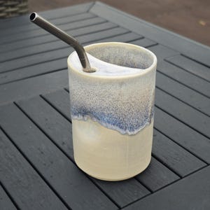 May include: A ceramic tumbler with a stainless steel straw. The tumbler has a cream-colored base with a blue and white speckled top. The straw is inserted into a hole on the side of the tumbler.