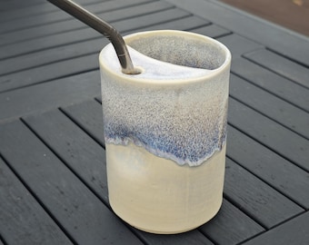 May include: A cylindrical ceramic tumbler with a cream-coloured base and a speckled blue and white glaze on the upper portion. The vessel features a built-in ceramic shelf near the rim with a circular opening holding a metallic drinking straw.