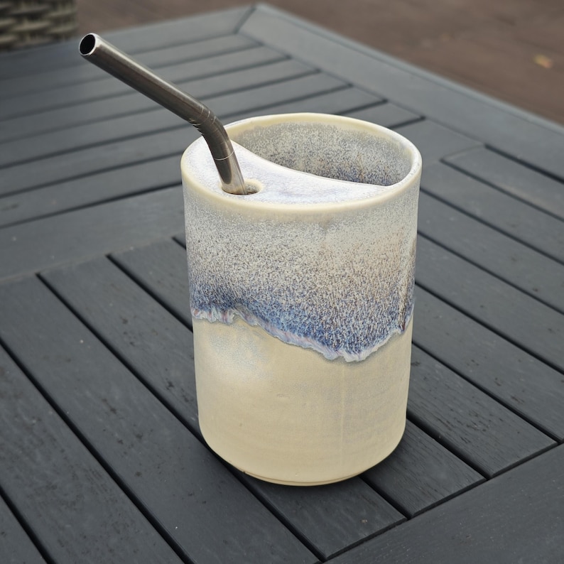 May include: A cylindrical ceramic tumbler with a cream-colored base and a speckled blue and white glaze on the upper portion. The vessel features a built-in ceramic shelf near the rim with a circular opening holding a metallic drinking straw.