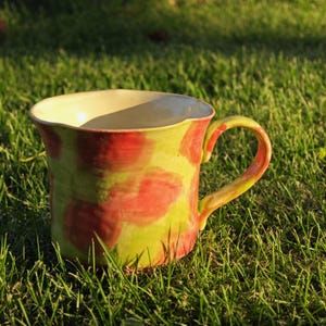 May include: A handmade ceramic mug with a vibrant design. The mug features a yellow interior and a green exterior adorned with large red splotches. It has a curved handle and sits on a bed of green grass.