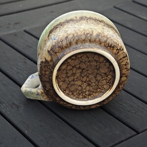May include: A ceramic mug with a textured, mottled brown and tan exterior. The rim and handle have a light blue and green design. The bottom of the mug is a darker brown with a textured pattern and a white rim.