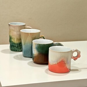 May include: Four ceramic mugs in a row, each with a unique design. The mugs feature a variety of colors, including green, blue, brown, and orange. Each mug has a handle and a white interior. The mugs are on a white surface.