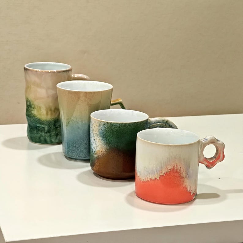 May include: Four ceramic mugs in a row, each with a unique design. The mugs feature a variety of colors, including green, blue, brown, and orange. Each mug has a handle and a white interior. The mugs are on a white surface.