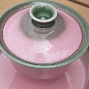 Puede incluir: Un juego de t&eacute; gaiwan de cer&aacute;mica que incluye un cuenco, una tapa y un platillo con un acabado de esmalte rosa brillante con efecto craquelado. El borde del cuenco, el borde del platillo y el pomo de la tapa tienen un esmalte verde salvia.