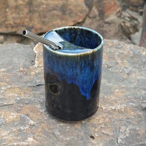 May include: A cylindrical ceramic tumbler featuring a glossy glaze that transitions from a deep black base to a vibrant cobalt blue rim. The vessel includes a built-in ceramic straw holder at the top edge and a metallic silver-toned straw.