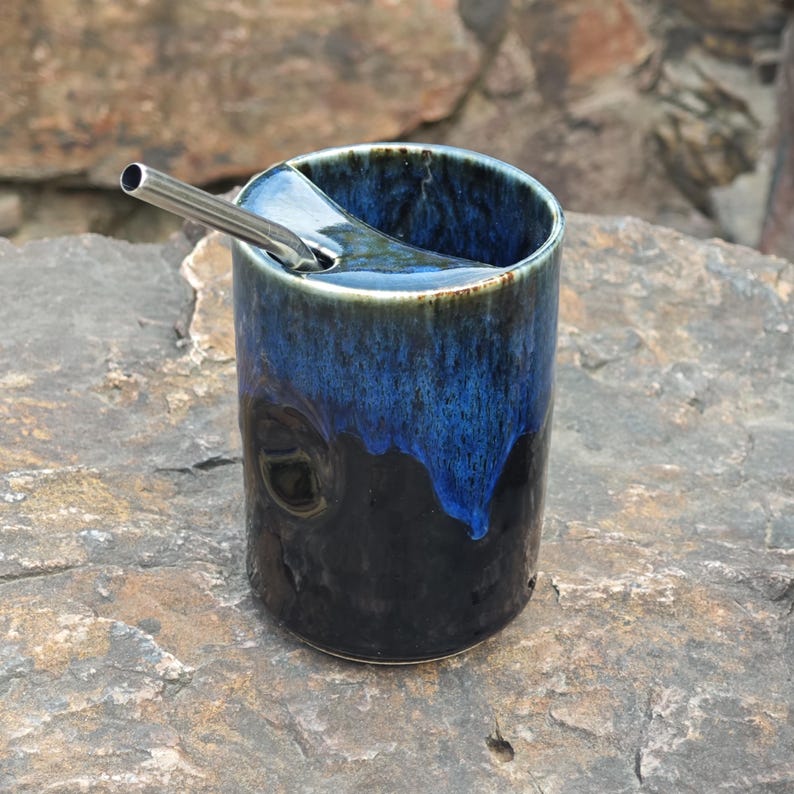 May include: A cylindrical ceramic tumbler featuring a glossy glaze that transitions from a deep black base to a vibrant cobalt blue rim. The vessel includes a built-in ceramic straw holder at the top edge and a metallic silver-toned straw.