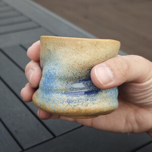 Peut inclure: Une tasse en c&eacute;ramique faite &agrave; la main avec une surface textur&eacute;e unique. La tasse pr&eacute;sente un m&eacute;lange de couleurs, notamment le beige, le bleu et le marron, cr&eacute;ant une esth&eacute;tique rustique. La tasse est tenue dans une main, mettant en valeur sa taille et sa forme.