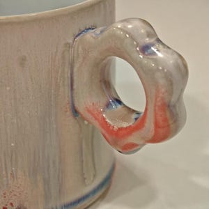 May include: Close-up of a ceramic mug with a unique handle design. The handle is a light color with red and blue accents. The mug has a light-colored body with blue stripes near the base.