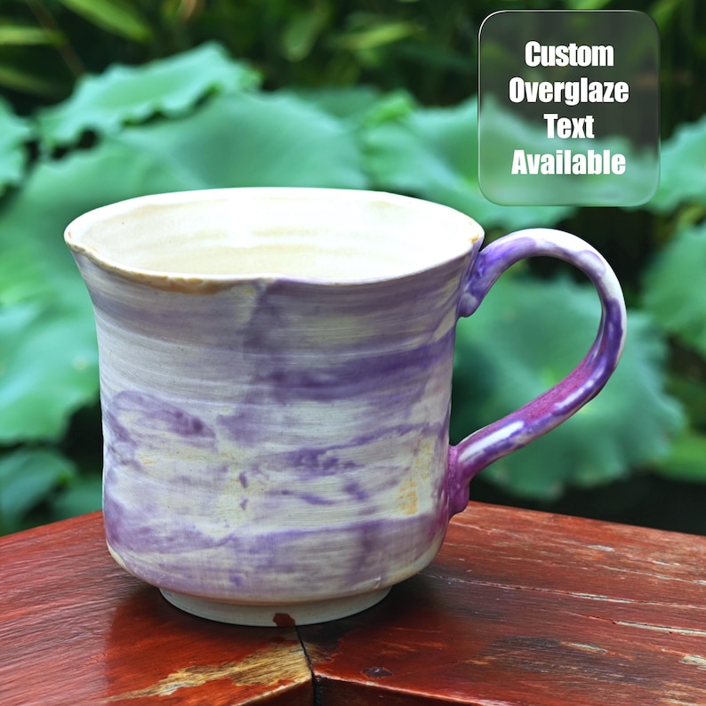 May include: A handmade ceramic mug with a white interior and a purple and white marbled exterior. The handle is purple. The mug has the text "Custom Overglaze Text Available".