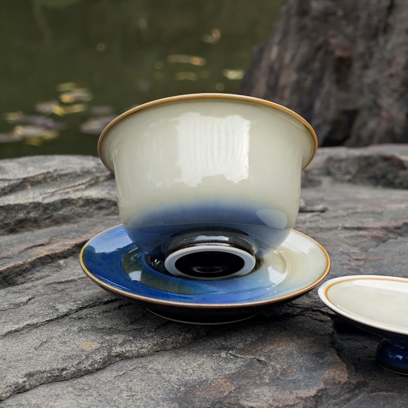 May include: A ceramic tea bowl with a lid, featuring a smooth cream-colored upper section that transitions into a deep blue base. The bowl rests on a matching saucer that displays a gradient of blue and cream tones with a dark, glossy rim.