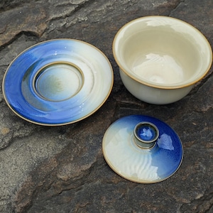 May include: A ceramic tea set consisting of a bowl, a saucer, and a lid. The bowl has a cream-colored interior and exterior. The saucer and lid feature a gradient design transitioning from cream to a deep blue, with a brown rim on all pieces.