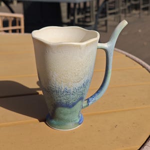 May include: A handmade ceramic mug with a unique handle design. The mug features a gradient of colors, starting with a creamy white at the top, transitioning to blue and green at the base. The handle is a curved, elongated shape, adding to its artistic appeal.