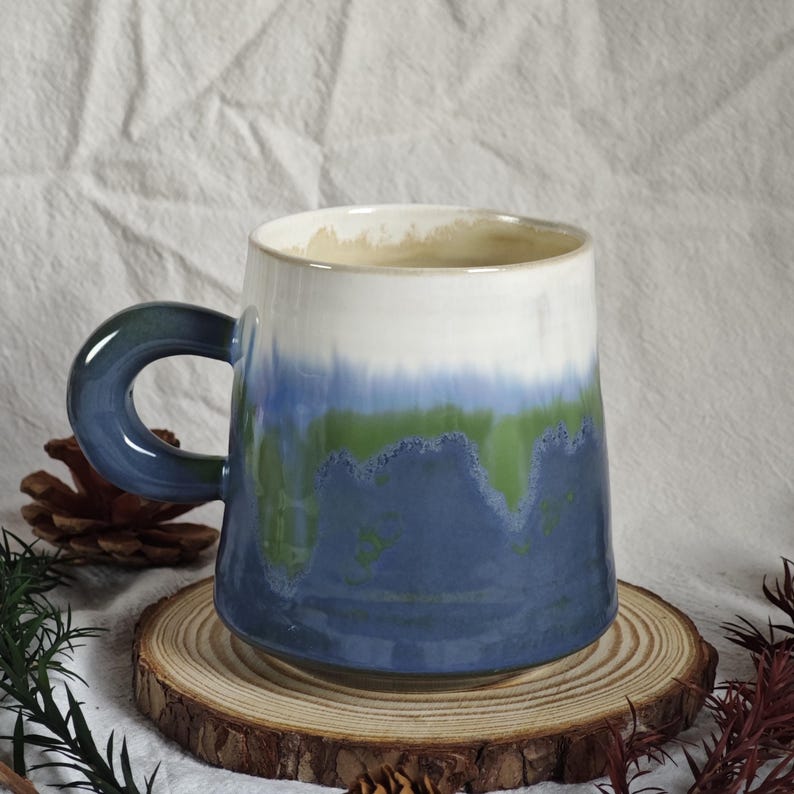 May include: A ceramic mug with a gradient design in shades of blue, green, and white. The mug has a rounded handle and sits on a wooden coaster. The mug's design resembles a landscape.
