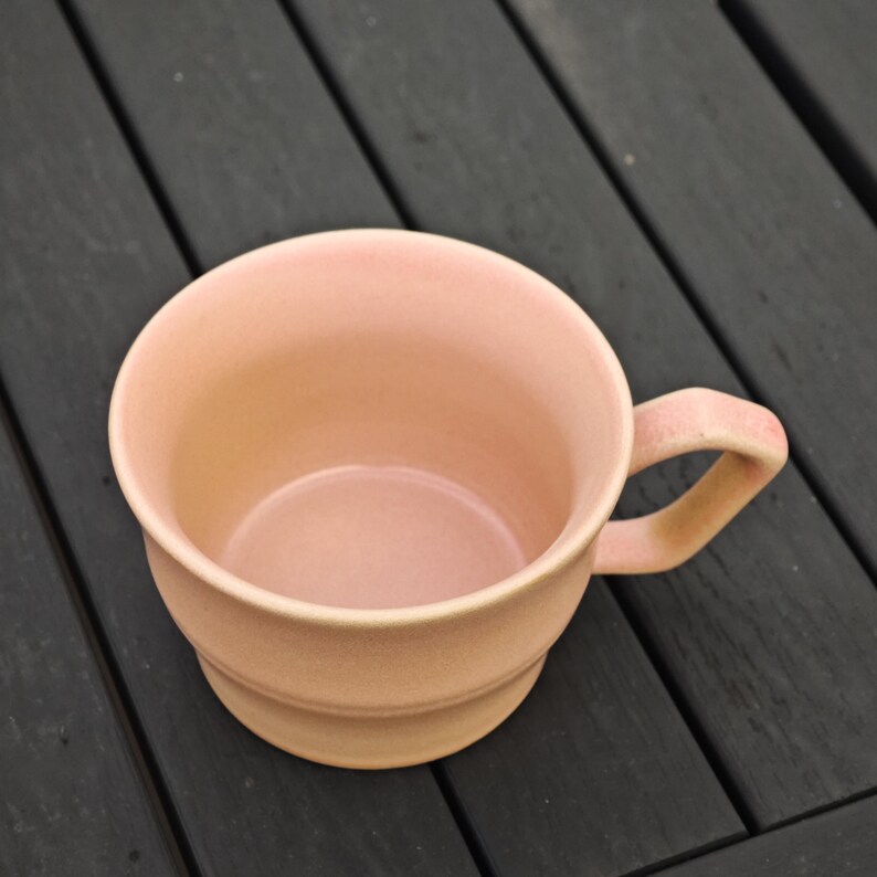 May include: A light pink ceramic mug with a rounded body and a handle. The mug has a slightly textured surface and a simple, classic design. The mug is empty and appears to be clean.